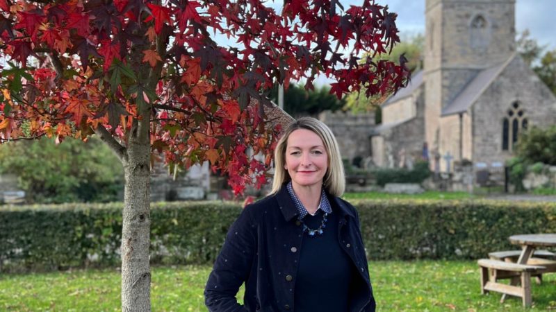 Jessica Morden MP stands in Redwick village hall. Also in the image is the historical church and an autumnal tree with vibrant red leaves.