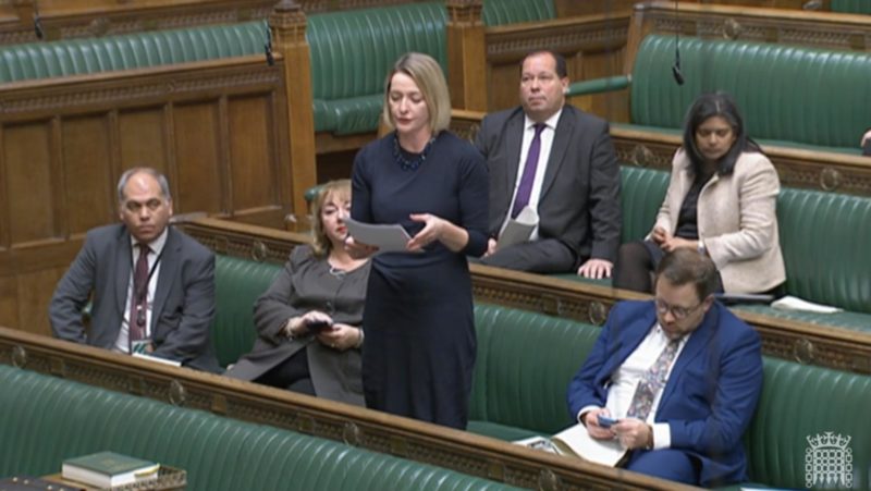Jessica Morden in the House of Commons asking a question to the Chancellor. Jessica is accompanied by other MPs. She is standing and wearing a blue dress. The green benches of Parliament are behind her. 