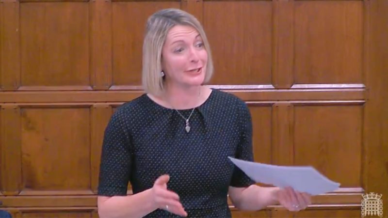 Jessica Morden MP in Parliament. Jessica is wearing a black dress and is stood against a wood-panelled wall.